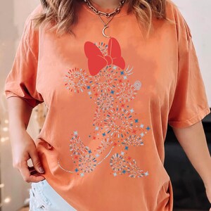 May include: Orange t-shirt featuring a Minnie Mouse design made of red, white, and blue fireworks. The shirt has a large red bow at the top and a relaxed fit. A fun and festive graphic tee.