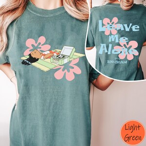 Puede incluir: Camiseta verde claro con un personaje de dibujos animados, un tocadiscos y flores rosas en la parte delantera. La parte trasera de la camiseta muestra el texto "Leave Me Alone" con flores rosas y el logo de Disney Lilo & Stitch.