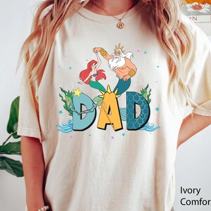 May include: Ivory-coloured t-shirt with a colourful graphic featuring Ariel, King Triton, and the word "DAD" in large letters. The design includes sea elements like waves and seaweed, with a star and sun graphic. The shirt is a Comfort Colors brand.