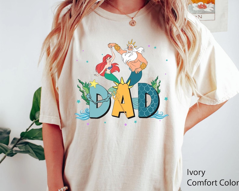 May include: Ivory-coloured t-shirt with a colourful graphic featuring Ariel, King Triton, and the word "DAD" in large letters. The design includes sea elements like waves and seaweed, with a star and sun graphic. The shirt is a Comfort Colors brand.