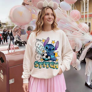 May include: Cream-colored sweatshirt featuring a graphic of the Disney character Stitch with a soccer ball and the word "Stitch". The design includes stars and the Disney logo. The person is wearing a pink pleated skirt.