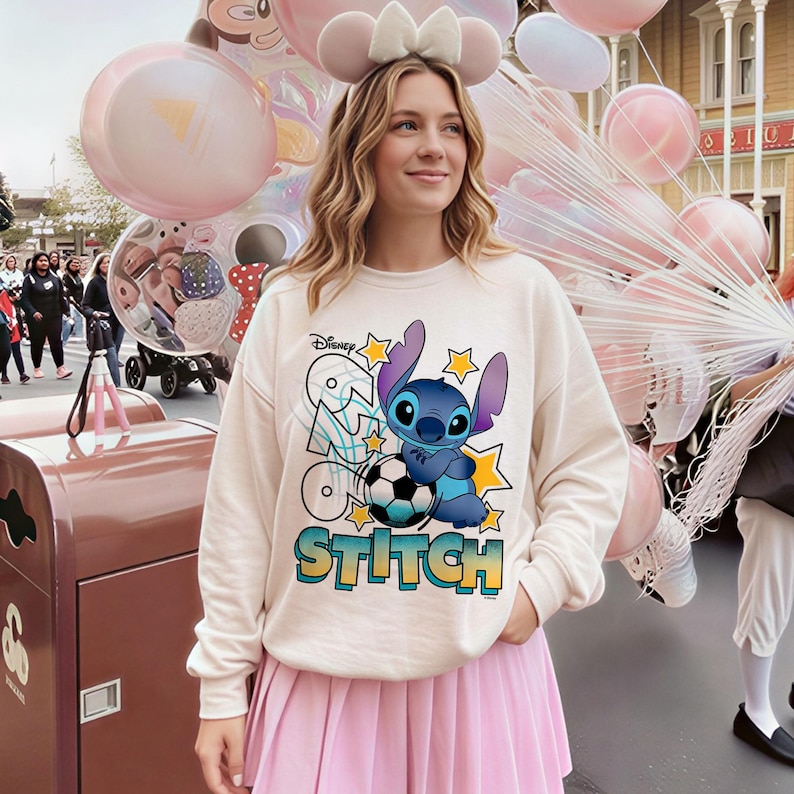 May include: Cream-colored sweatshirt featuring a graphic of the Disney character Stitch with a soccer ball and the word "Stitch". The design includes stars and the Disney logo. The person is wearing a pink pleated skirt.