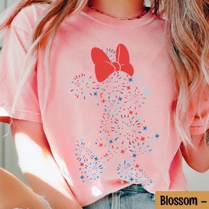 May include: A light pink t-shirt featuring a red bow and a design of red, white, and blue fireworks. The shirt has a relaxed fit and short sleeves. The word "Blossom" is printed on the bottom right.