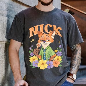 May include: Dark grey t-shirt with a cartoon fox character named Nick, surrounded by flowers. The word "NICK" is arched above the fox in orange letters. The fox holds a paw-shaped lollipop. The shirt has a vintage, worn appearance.