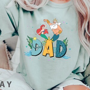 May include: A pale green t-shirt with a vibrant graphic of Ariel and King Triton dancing above the word "DAD". The design incorporates aquatic elements such as seaweed, stars, and waves, creating a whimsical, underwater theme.