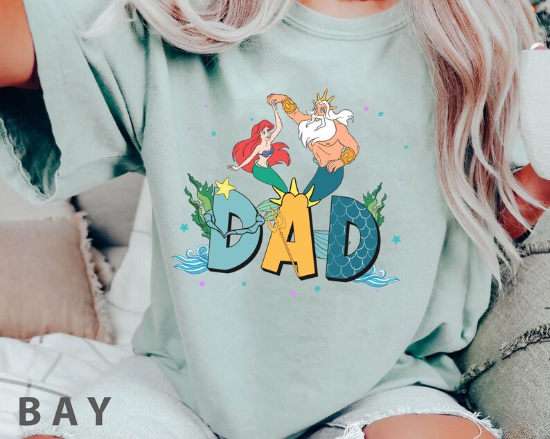 May include: A pale green t-shirt with a vibrant graphic of Ariel and King Triton dancing above the word "DAD". The design incorporates aquatic elements such as seaweed, stars, and waves, creating a whimsical, underwater theme.