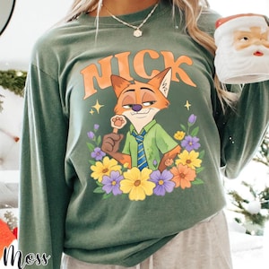 May include: Green long-sleeve shirt with a cartoon fox character named Nick, surrounded by flowers. The word "NICK" is printed above the fox in orange letters. The shirt has a relaxed fit.