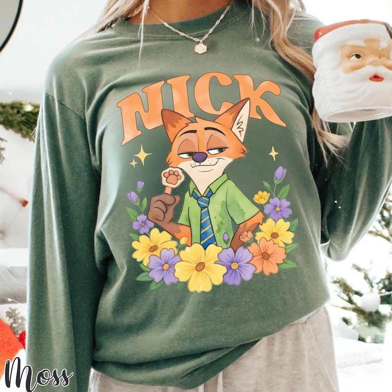 May include: Green long-sleeve shirt with a cartoon fox character named Nick, surrounded by flowers. The word "NICK" is printed above the fox in orange letters. The shirt has a relaxed fit.