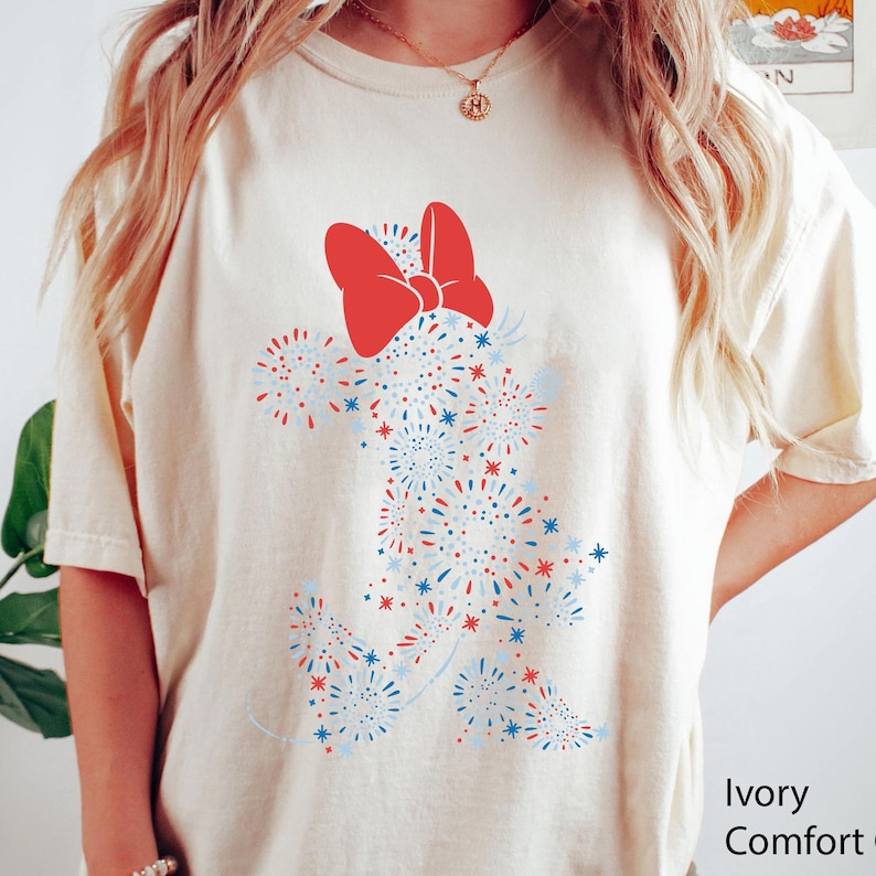 May include: Ivory-colored t-shirt featuring a Minnie Mouse design made of red, white, and blue fireworks. The design includes a large red bow at the top. The text "Ivory Comfort" is visible on the bottom right.