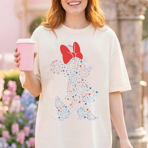 May include: Cream-colored t-shirt featuring a Minnie Mouse design made of red, white, and blue fireworks. The design includes a large red bow. The shirt is a relaxed fit.