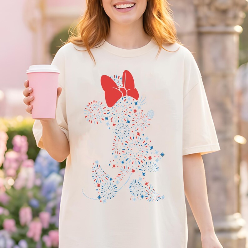 May include: Cream-colored t-shirt featuring a Minnie Mouse design made of red, white, and blue fireworks. The design includes a large red bow. The shirt is a relaxed fit.