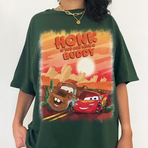 May include: Dark green t-shirt featuring a graphic of the characters Mater and Lightning McQueen from the movie Cars, with the text "HONK IF YOU ARE WITH A BUDDY" in orange and red. The background depicts a desert landscape with a sunset.