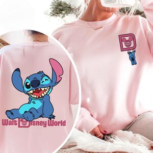 May include: A light pink sweatshirt featuring a cartoon Stitch design. The front has a small Stitch graphic hanging from a pink square with a Mickey Mouse head. The back displays a larger Stitch graphic with the text "Walt Disney World".