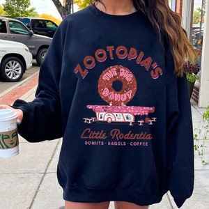 Puede incluir: Sudadera azul marino con un gr&aacute;fico que presenta el texto "ZOOTOPIA'S THE BIG DONUT" sobre una rosquilla rosa y un cami&oacute;n de comida rosa. Debajo del cami&oacute;n est&aacute; el texto "Little Rodentia: Donuts, Bagels, Coffee."