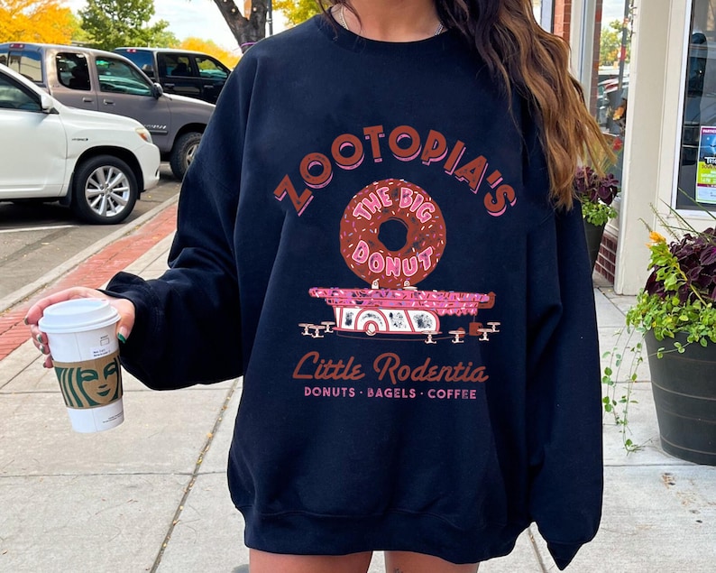 Puede incluir: Sudadera azul marino con un gr&aacute;fico que presenta el texto "ZOOTOPIA'S THE BIG DONUT" sobre una rosquilla rosa y un cami&oacute;n de comida rosa. Debajo del cami&oacute;n est&aacute; el texto "Little Rodentia: Donuts, Bagels, Coffee."