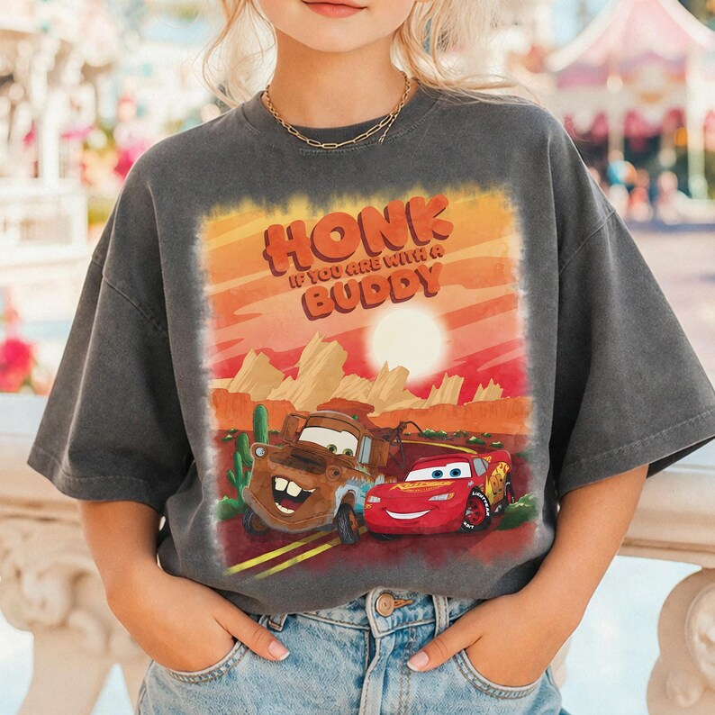 May include: Dark gray t-shirt featuring a graphic of two cartoon cars with the text "HONK IF YOU ARE WITH A BUDDY" in orange and red. The design includes a desert landscape with a setting sun.