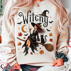 May include: A cream-colored sweatshirt featuring a graphic print of a person in a black dress and hat sitting on a broomstick, holding a glass. The design includes a black cat, a crescent moon, bats, and orange and yellow autumn leaves.