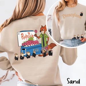 Puede incluir: Sudadera de color arena con una ilustraci&oacute;n de dibujos animados de Nick Wilde vendiendo Pawpsicles. El dise&ntilde;o incluye el texto "Pawpsicle" y varios personajes peque&ntilde;os. El lado derecho de la sudadera tiene la palabra "Disneyland" y un personaje peque&ntilde;o.