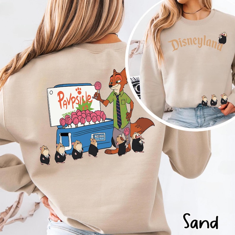 Puede incluir: Sudadera de color arena con una ilustraci&oacute;n de dibujos animados de Nick Wilde vendiendo Pawpsicles. El dise&ntilde;o incluye el texto "Pawpsicle" y varios personajes peque&ntilde;os. El lado derecho de la sudadera tiene la palabra "Disneyland" y un personaje peque&ntilde;o.