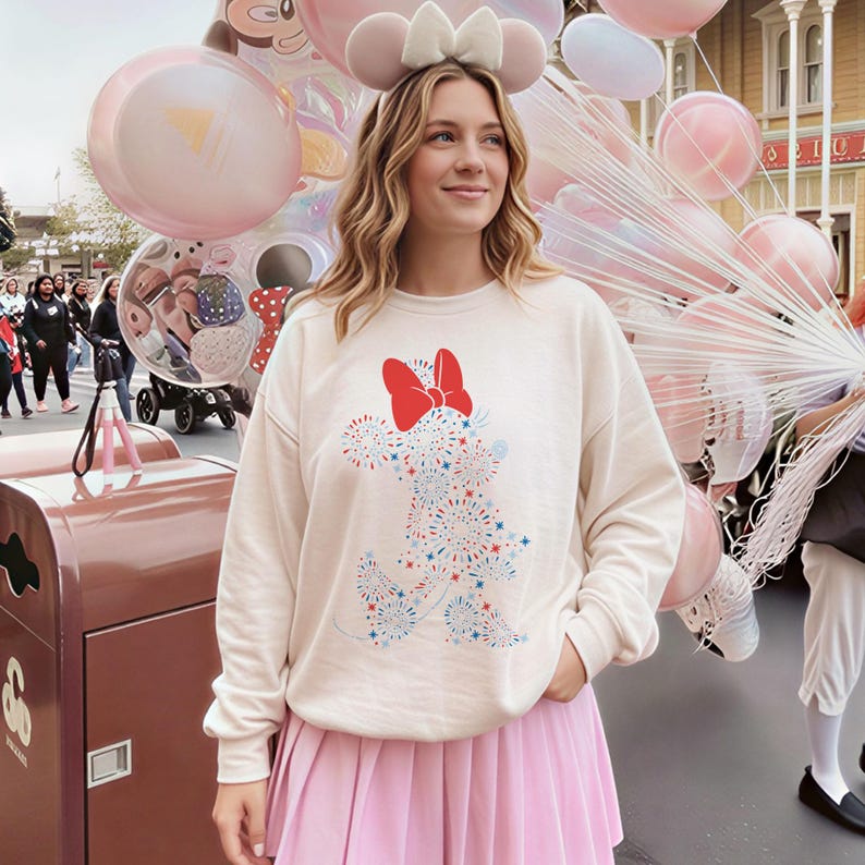 May include: Cream-colored sweatshirt featuring a Minnie Mouse design made of red, white, and blue fireworks. The sweatshirt has a red bow at the top. The person is wearing a pink pleated skirt and a Minnie Mouse ear headband. Pink balloons are in the background.