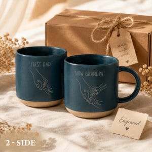 May include: Two teal ceramic mugs with a tan base. One mug reads "FIRST DAD" and the other reads "NOW GRANDPA". Both mugs feature a hand-holding design with names. A gift box and tag that says "MADE with LOVE" are in the background.