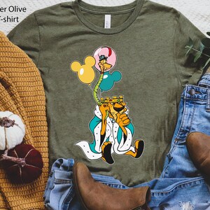 May include: A green Bella T-shirt with a cartoon image of a lion wearing a crown and holding balloons. The balloons are yellow, blue, and pink. The lion is wearing a blue and white robe. The shirt is heather olive.