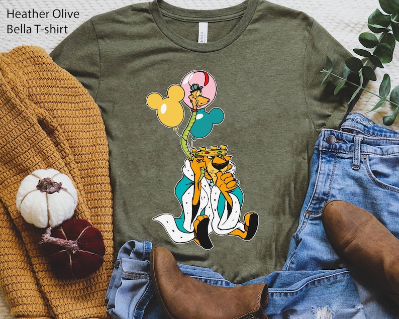 May include: A green Bella T-shirt with a cartoon image of a lion wearing a crown and holding balloons. The balloons are yellow, blue, and pink. The lion is wearing a blue and white robe. The shirt is heather olive.