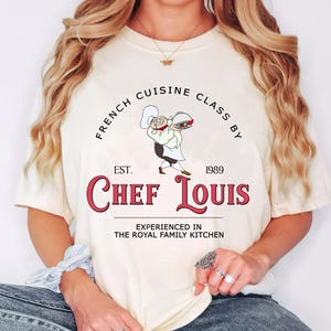 May include: Cream-colored t-shirt featuring a cartoon chef holding a platter. The shirt has the text "FRENCH CUISINE CLASS BY" above "CHEF LOUIS" in red, with "EST. 1989" and "EXPERIENCED IN THE ROYAL FAMILY KITCHEN" below.