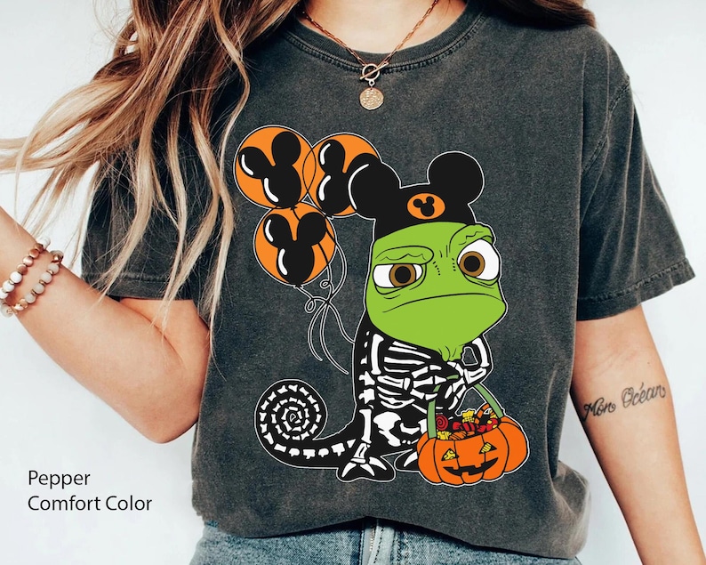 May include: A charcoal grey t-shirt featuring a graphic of a green chameleon wearing a black skeleton costume and a black mouse-ear hat. The chameleon holds a pumpkin-shaped bucket filled with sweets and three orange mouse-ear balloons.