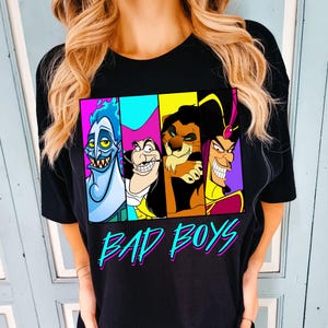 May include: Black t-shirt featuring a colorful graphic of four cartoon villains. The graphic includes Hades, Captain Hook, Scar, and Jafar, with the words "BAD BOYS" in a teal and pink font. The shirt is a crew neck style.