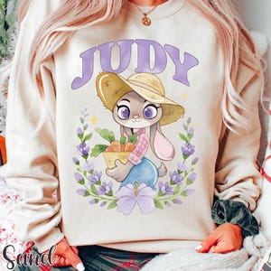 May include: Cream-coloured sweatshirt with a cartoon bunny wearing a straw hat and holding a basket of carrots, surrounded by a floral wreath. The name "JUDY" is arched above the bunny in purple letters. The sweatshirt has long sleeves.