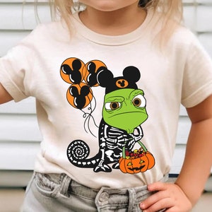 May include: A cream-coloured t-shirt featuring a graphic of a green chameleon wearing a black Mickey Mouse ear hat, holding orange balloons with Mickey Mouse silhouettes, and carrying a pumpkin bucket filled with colourful candy.