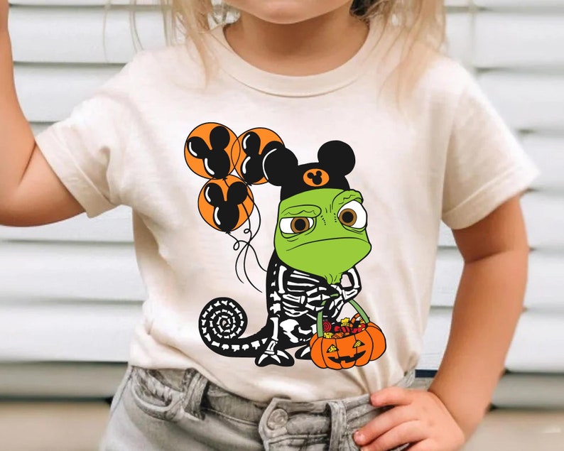 May include: A cream-coloured t-shirt featuring a graphic of a green chameleon wearing a black Mickey Mouse ear hat, holding orange balloons with Mickey Mouse silhouettes, and carrying a pumpkin bucket filled with colourful candy.