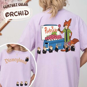 Puede incluir: Una camiseta de color lila claro con la palabra "Disneyland" en escritura dorada en la parte delantera. La parte trasera presenta un zorro de dibujos animados vendiendo paletas de un refrigerador, con el texto "Pavpsicle". La camiseta est&aacute; etiquetada como "Comfort Color Orchid".