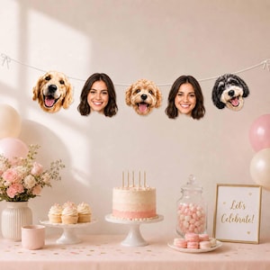 May include: A party banner featuring five cutouts of faces, including three dogs and two women, strung on a white cord. The display includes a tiered cake, cupcakes, a jar of pink candies, a vase of flowers, and a sign reading Let's Celebrate.