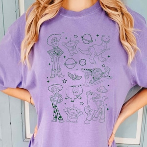 Puede incluir: Una camiseta morada con dibujos lineales verdes de personajes de Toy Story. El dise&ntilde;o incluye a Woody, Buzz Lightyear, extraterrestres, dinosaurios y otros personajes. La camiseta es de color liso y tiene cuello redondo.