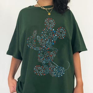 May include: Dark green t-shirt featuring a graphic of Mickey Mouse made of red, white, and blue fireworks. The shirt has short sleeves and a relaxed fit. A gold necklace is visible at the neckline.