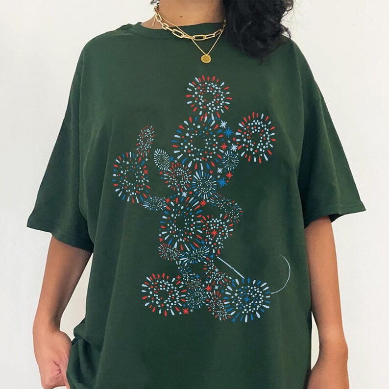 May include: Dark green t-shirt featuring a graphic of Mickey Mouse made of red, white, and blue fireworks. The shirt has short sleeves and a relaxed fit. A gold necklace is visible at the neckline.