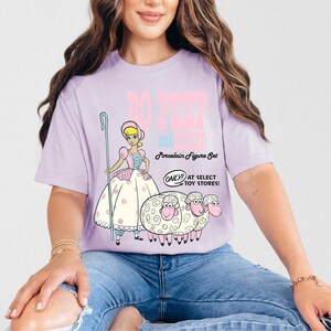 May include: A purple t-shirt with a graphic of Bo Peep and her sheep from the Toy Story movie. The text on the shirt reads "Bo Peep and her sheep Porcelain Figure Set Only at Select Toy Stores!"