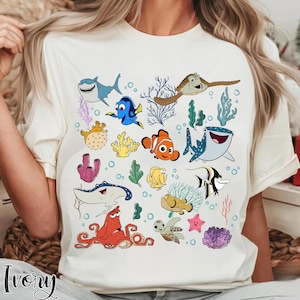 Può includere: T-shirt color avorio con un colorato design a cartoni animati di varie creature marine, tra cui uno squalo, un pesce pagliaccio e una tartaruga marina. Il design include elementi di corallo e alghe, creando una scena sottomarina. La parola "Ivory" è stampata in basso.