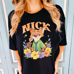 May include: Black t-shirt with a cartoon fox character named Nick, wearing a green shirt and blue tie, surrounded by colourful flowers. The word "NICK" is arched above the fox in orange letters. The design includes stars and a paw-shaped lollipop.