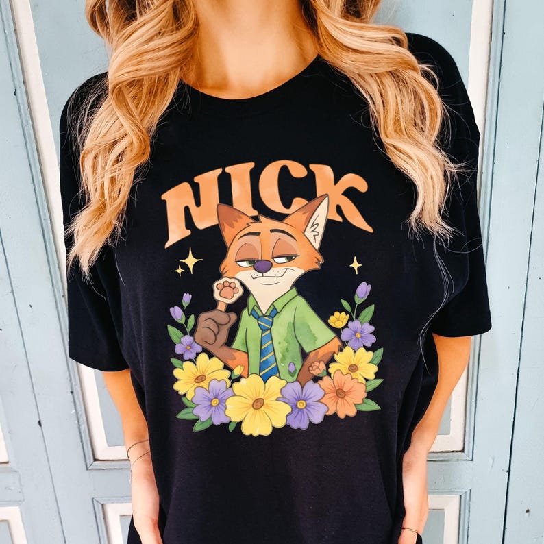 May include: Black t-shirt with a cartoon fox character named Nick, wearing a green shirt and blue tie, surrounded by colourful flowers. The word "NICK" is arched above the fox in orange letters. The design includes stars and a paw-shaped lollipop.