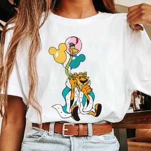 May include: White t-shirt with a cartoon illustration of a lion wearing a crown and holding balloons. The lion is wearing a blue and white robe. The balloons are yellow, blue, and pink.