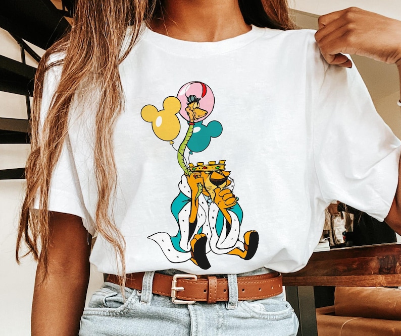 May include: White t-shirt with a cartoon illustration of a lion wearing a crown and holding balloons. The lion is wearing a blue and white robe. The balloons are yellow, blue, and pink.