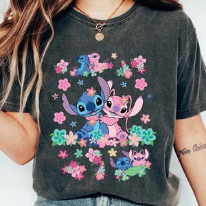 May include: Dark gray t-shirt featuring a colorful graphic of the characters Stitch and Angel from Lilo & Stitch, surrounded by pink and teal flowers. The design includes multiple images of the characters. The shirt is a casual wear item.