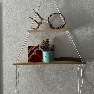 May include: Two wooden shelves hanging from the ceiling with white rope. The top shelf has a white antler and a brown wreath. The bottom shelf has a colorful box, a red heart, a blue and white can, and a wooden sign that says "Boxy Eat".