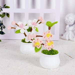 Op de afbeelding: Twee kleine witte keramische potten met elk een gehaakt bloemstuk. De bloemen zijn roze, wit en geel, met groene bladeren en bruine stelen. De potten staan op een wit oppervlak.