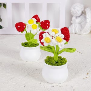 May include: Two small white ceramic pots each hold a crocheted flower arrangement. The flowers have white petals with yellow centers, green stems and leaves, and red strawberry accents. The pots sit on a textured white surface.