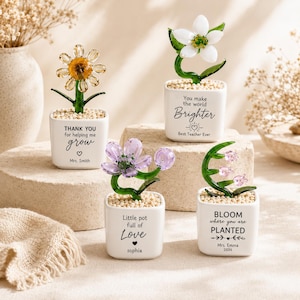 Può includere: Quattro piccoli vasi quadrati in ceramica bianca contenenti fiori in vetro con steli verdi e perline trasparenti. I vasi presentano scritte nere: Thank you for helping me grow, You make the world Brighter, Little pot full of Love e BLOOM where you are PLANTED.