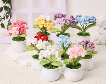 以下が含まれることがあります： 小さな白い鉢に入った手作りのかぎ針編みの花のコレクション。花はピンク、黄色、赤、白、青、紫など様々な色があり、緑の葉が付いています。鉢は白い表面に配置されています。